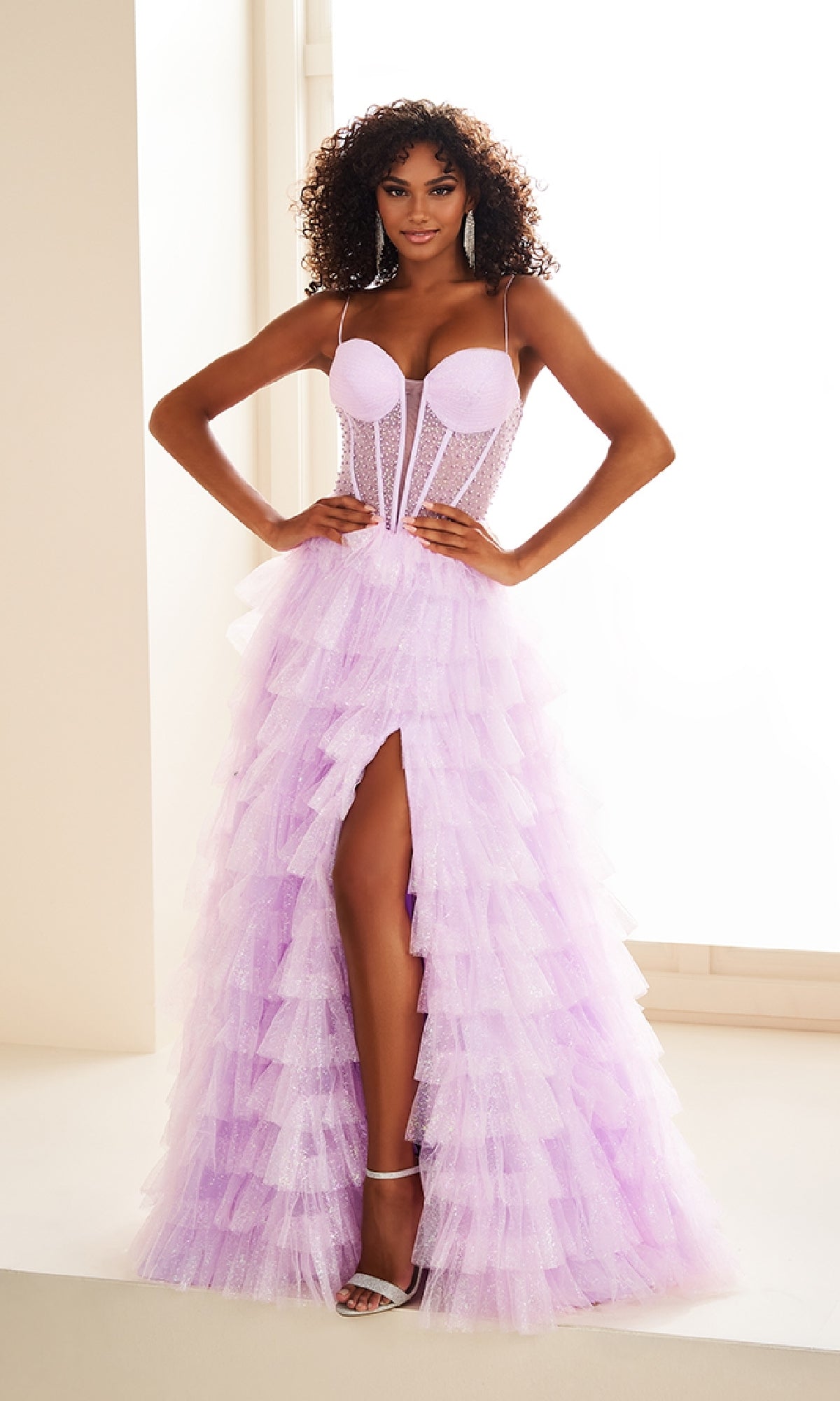 A woman stands indoors wearing the Ellie Wilde EW36029 Ruffled Prom Ball Gown, which features a light purple tiered ruffle tulle skirt with a high slit and spaghetti straps, paired with white high-heeled sandals.